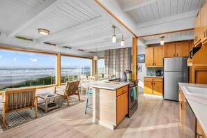A modern kitchen with wooden cabinets, a stainless steel refrigerator, and a large island. There is a view of the ocean through large windows. - Magnificent Oceanfront Retreat, Pet Friendly, Single Level, Deck and Fire-Pit w/ Views, EV Charging (Rockaway Beach)