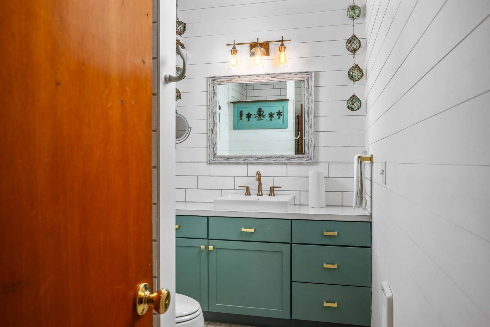 Un baño con paredes de azulejos blancos, un mueble de tocador verde con tiradores dorados, un lavabo blanco y un espejo con marco decorativo.