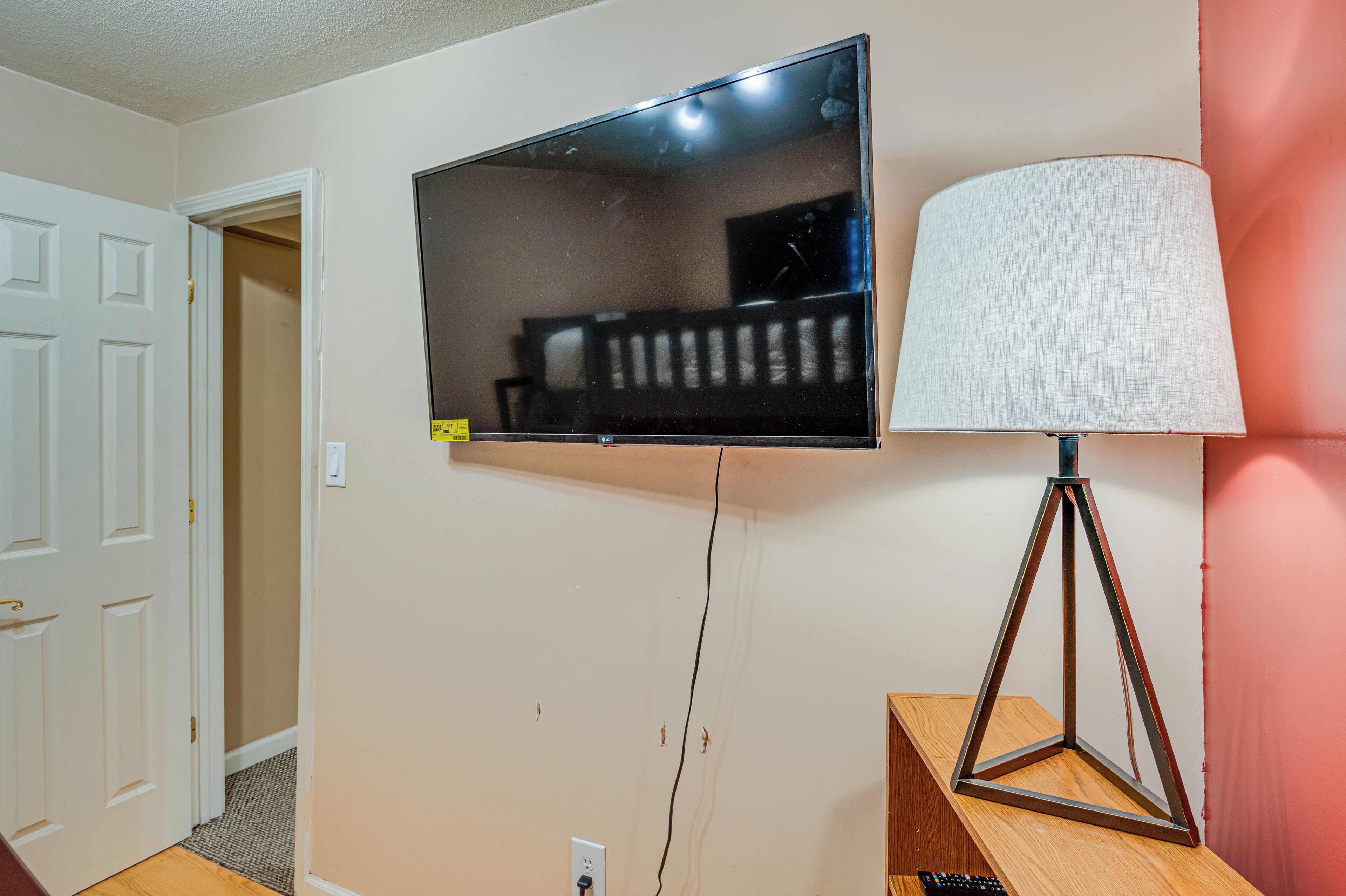Un televisor de pantalla plana montado en la pared, una lámpara sobre un soporte de madera y una puerta blanca.