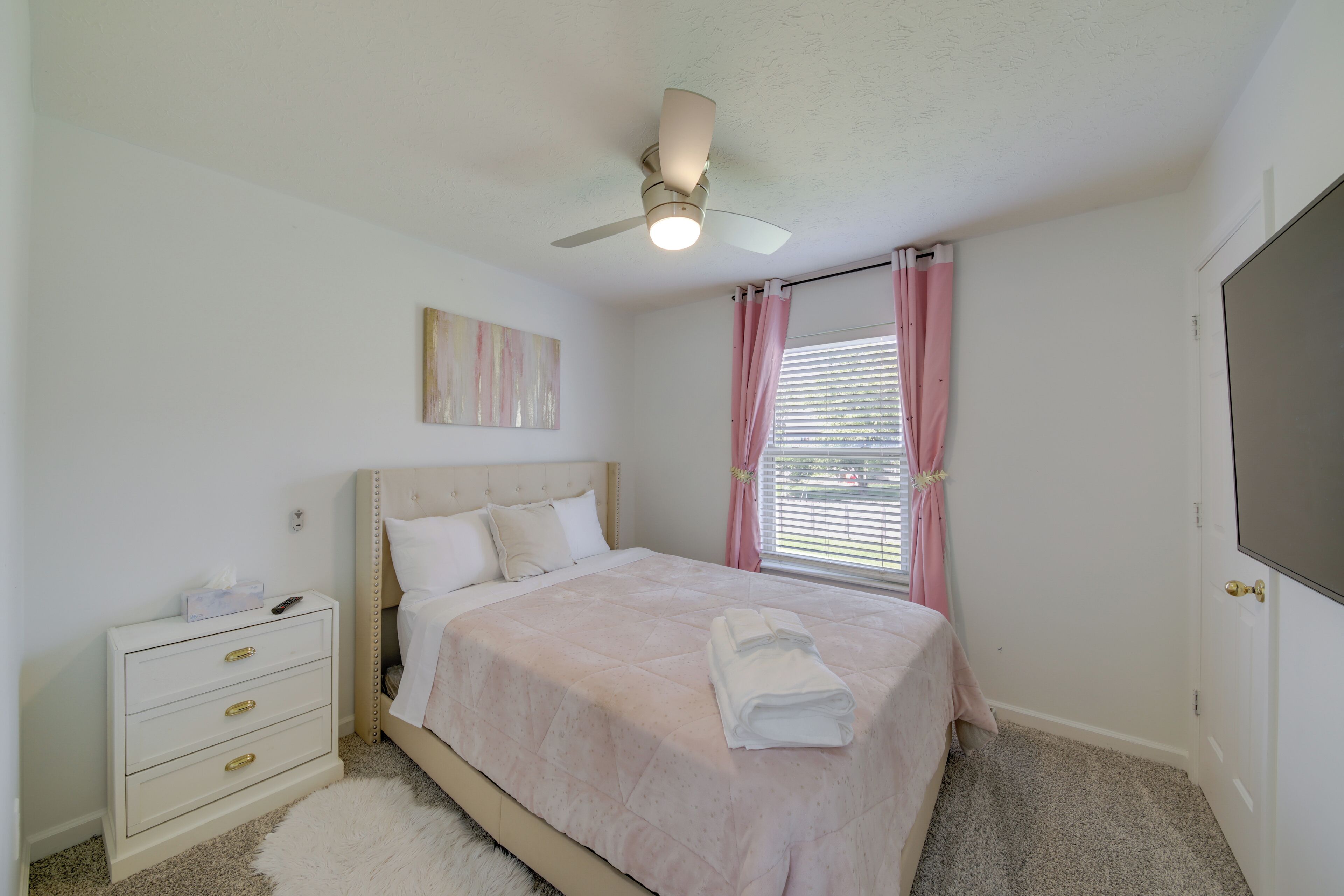 A bedroom with a bed, a nightstand, a ceiling fan, a window with curtains, and a door.