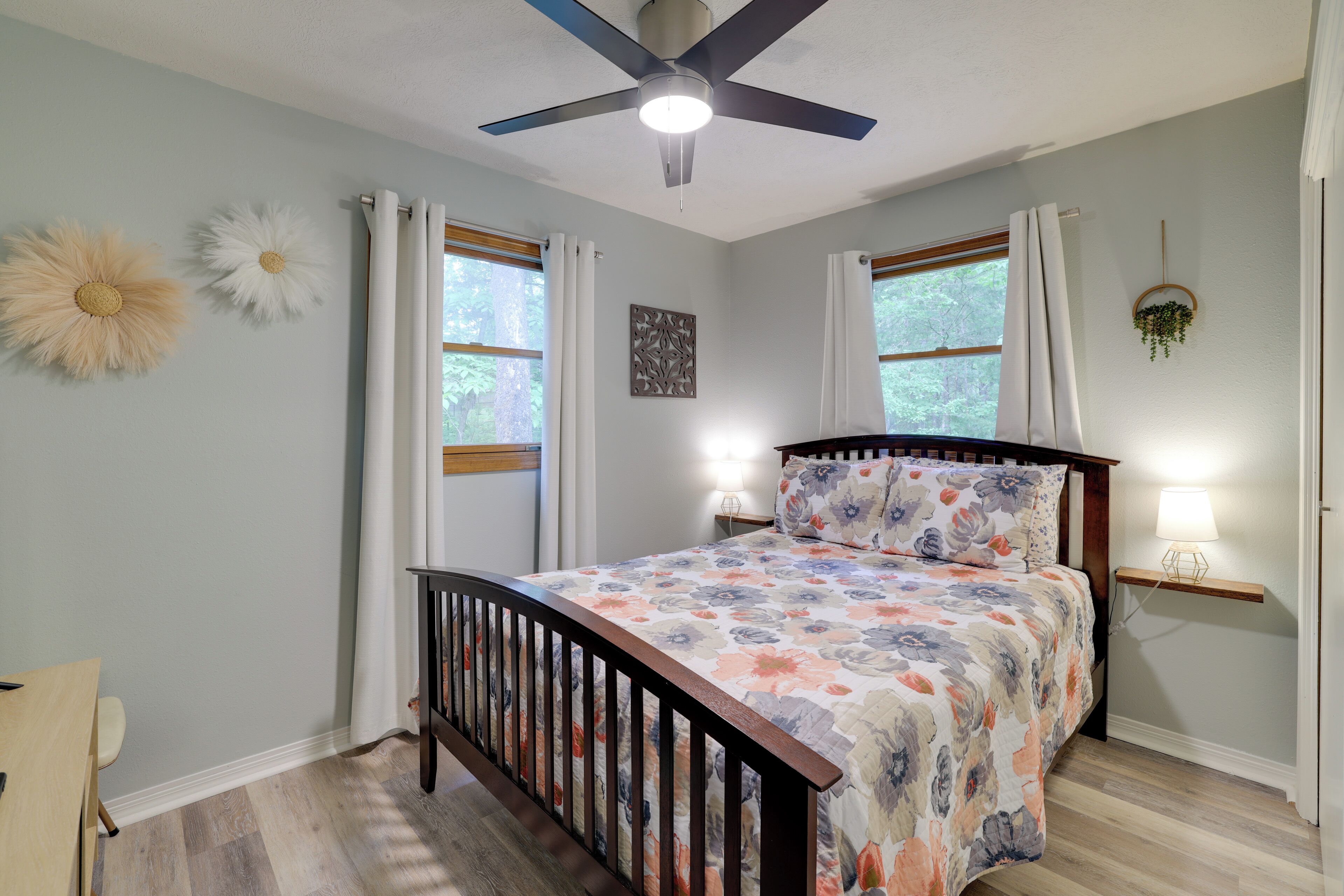 A bedroom with a bed, a ceiling fan, two windows with curtains, a bedside table with a lamp, and a wall-mounted plant.