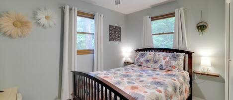 A bedroom with a bed, a ceiling fan, two windows with curtains, a bedside table with a lamp, and a wall-mounted plant.