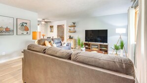 A living room with a brown leather sofa, a flat-screen TV, and a coffee table. There are decorative items on shelves and a potted plant on a side table. - Hot Tub & Fire Pit: Holiday Island Getaway! (Holiday Island)