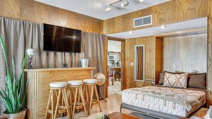 Ein modernes Hotelzimmer mit einer Wandpaneele aus Holz, einem Flachbildfernseher, einer Bar mit Hockern, einem Bett mit gemusterten Bettwäsche und einer Zimmerpflanze.