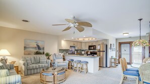 A spacious living room with a central island, a kitchen with modern appliances, and a dining area with wicker chairs.
