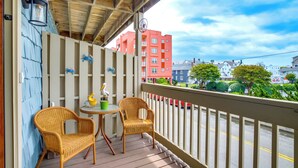A wooden balcony with wicker chairs and a small table, overlooking a street with buildings and greenery.