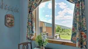 A room with a wooden table, a chair, a vase with flowers, a 'Welcome Friends' sign, and a view of a green landscape through a window. - Hike, Fish & Unwind! Catskills Studio w/ Mtn Views (Stamford)