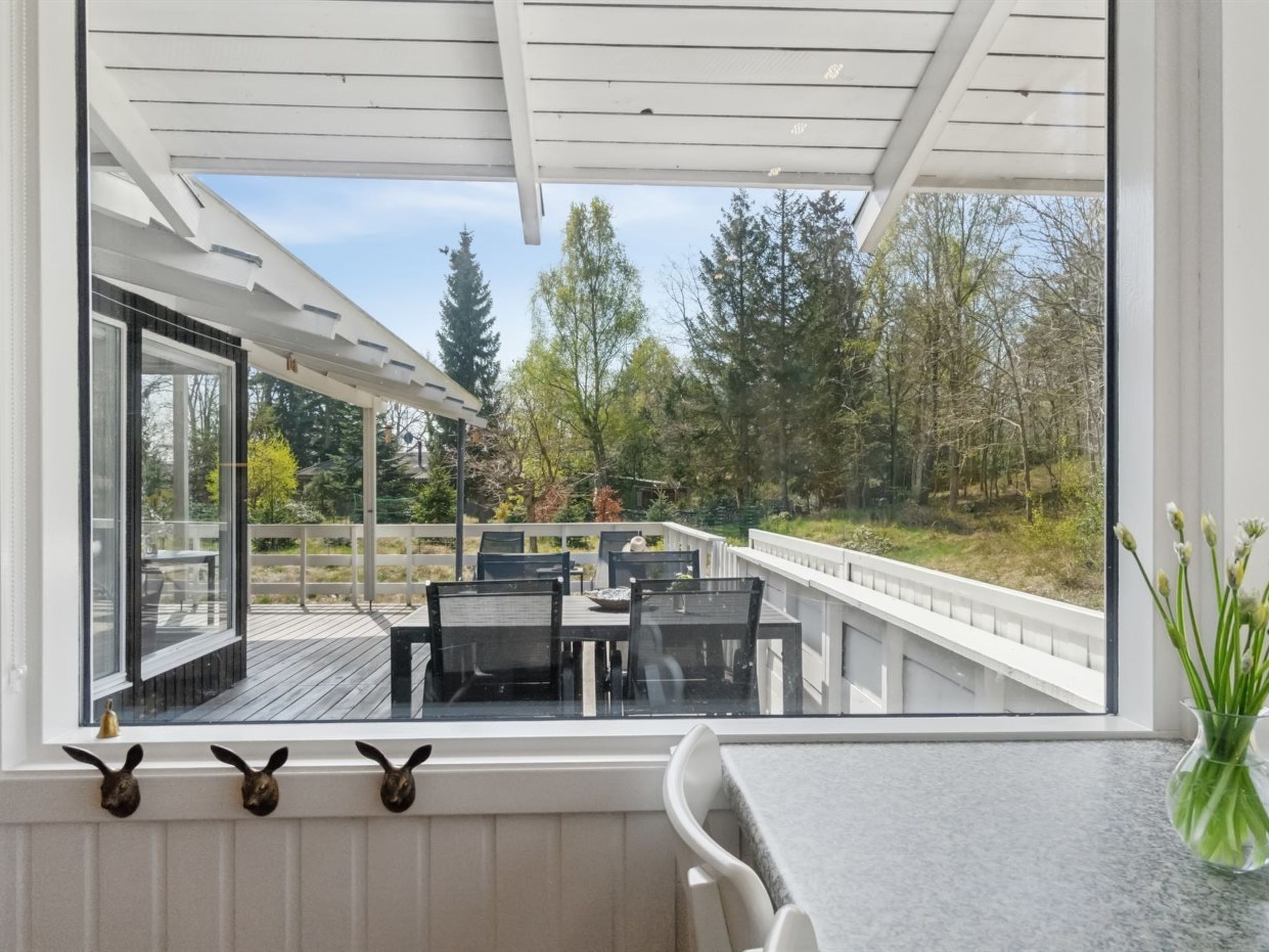 A modern outdoor deck with a glass railing, a dining area with chairs and a table, and a view of a forested area.