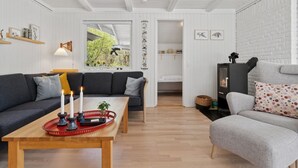 Een gezellige woonkamer met een houten tafel, een grijze bank en een grijze fauteuil. Er is een open haard en een raam met uitzicht op groen.