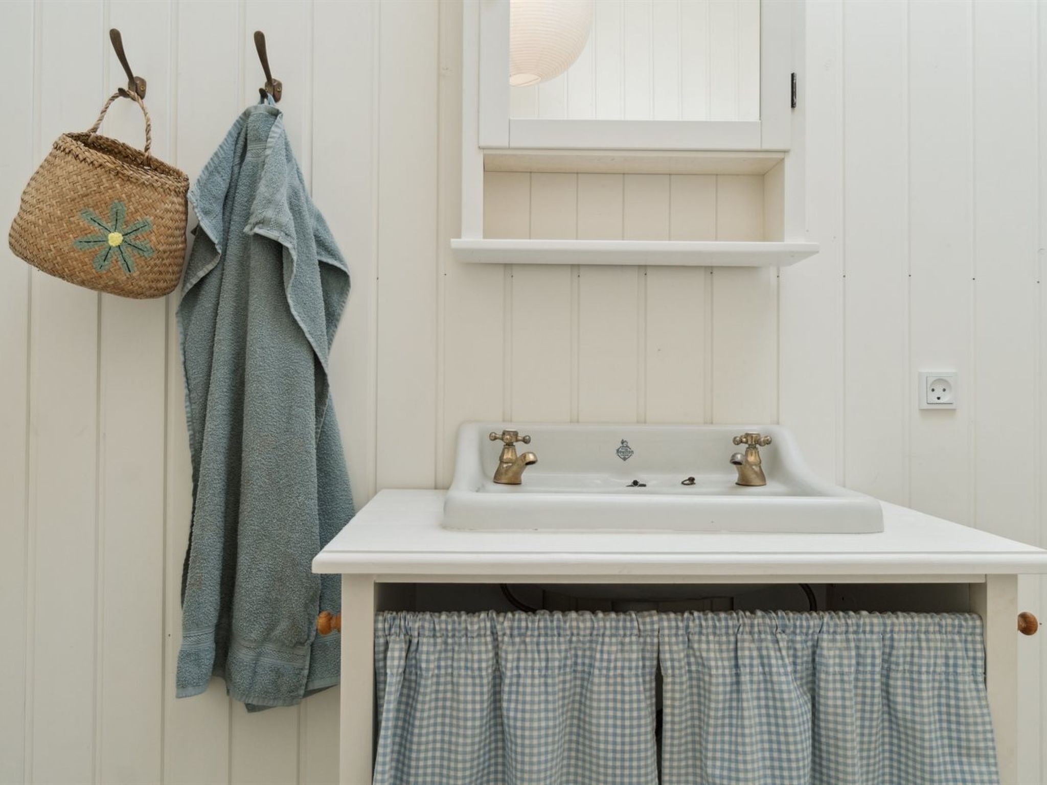 A bathroom with a white sink, two faucets, a mirror above the sink, and a hanging basket with a towel.