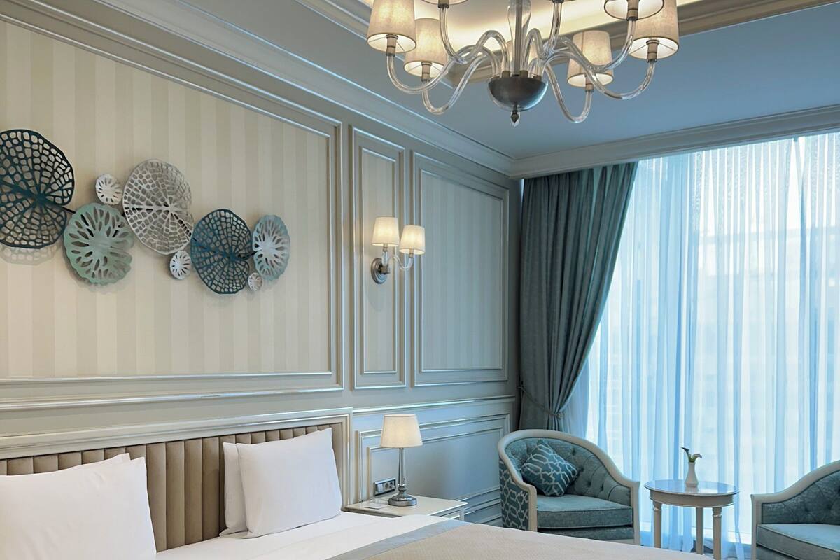 A hotel room with a large bed, a chandelier, decorative wall panels, and a view of the outdoors through sheer curtains.