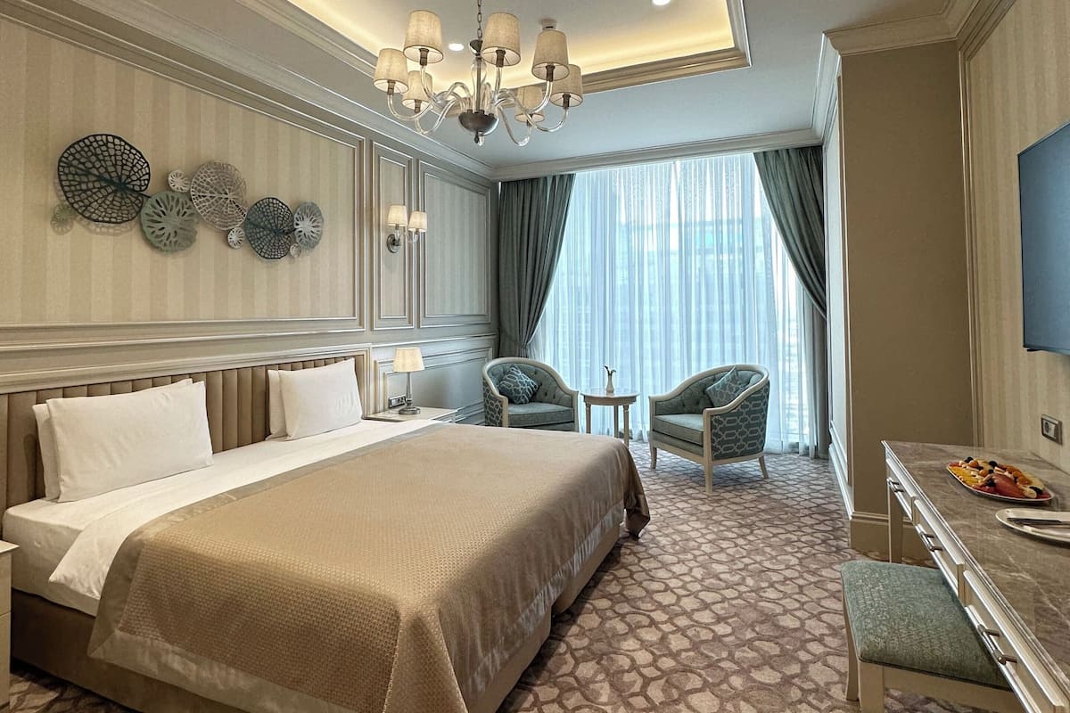 A hotel room with a large bed, a desk with a chair, a chandelier, and a view of the outdoors.
