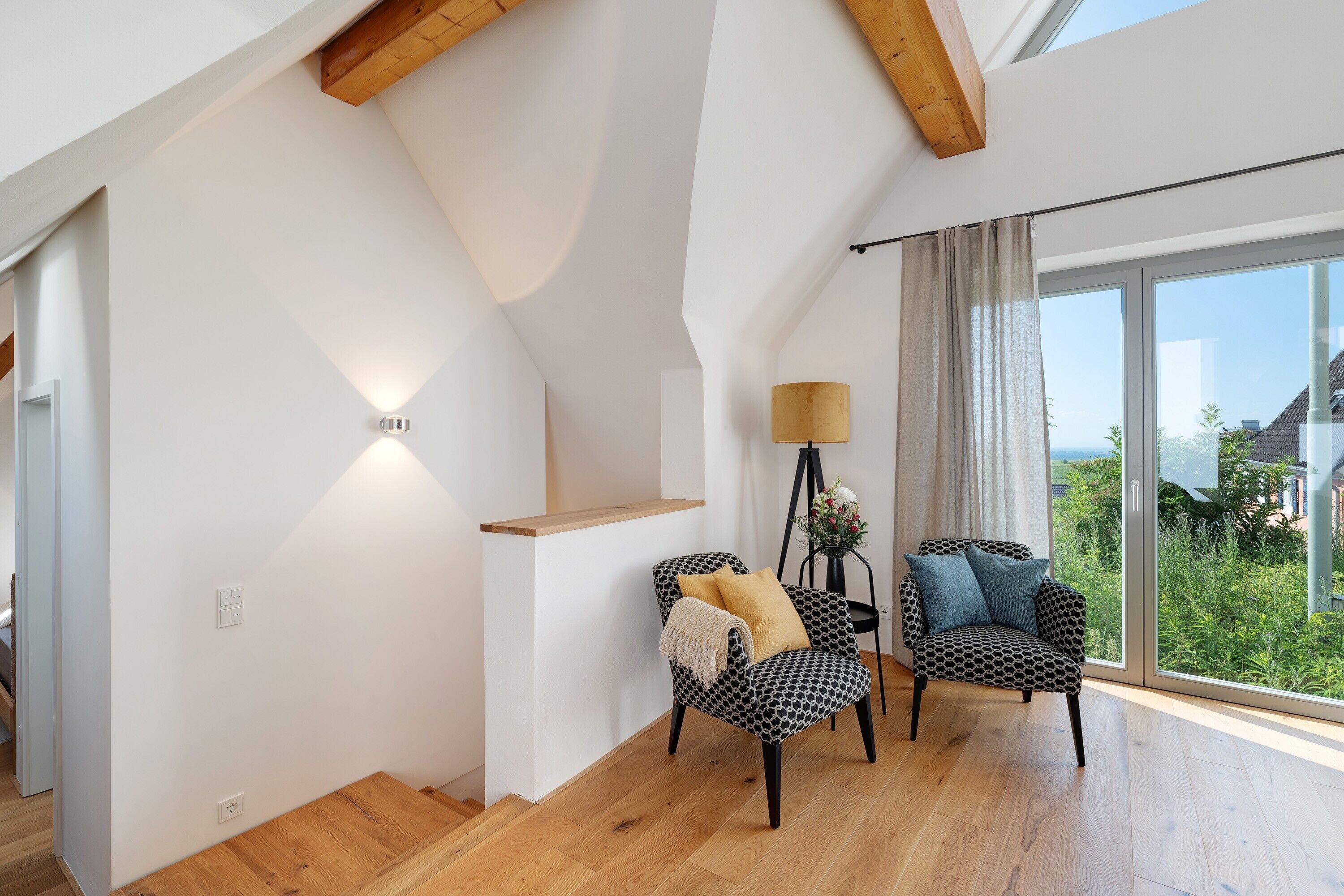 Ein modernes Interieur mit hoher Decke, Holzbalken, einer Treppe und Blick durch große Fenster.