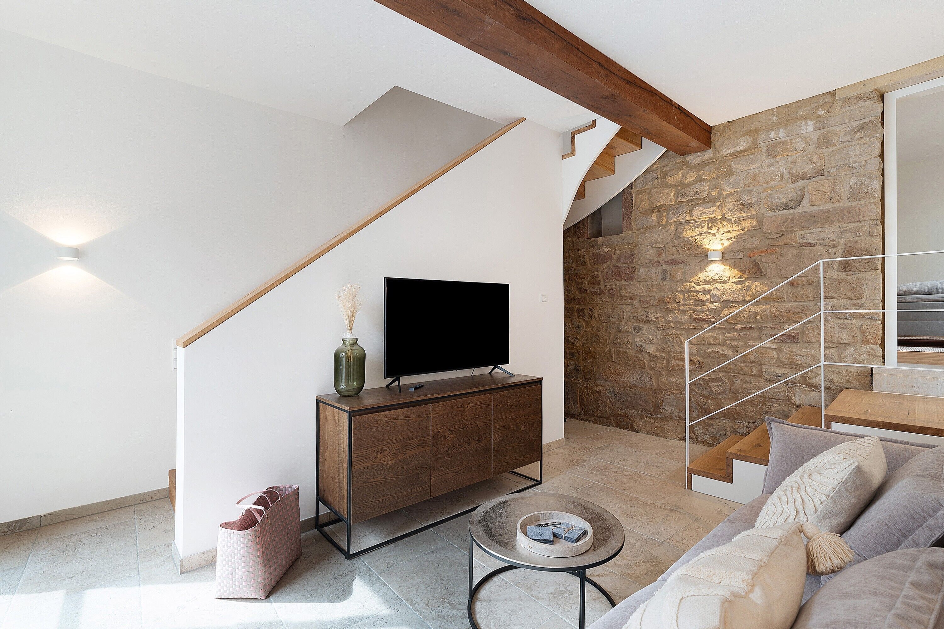 Un salon moderne doté d’un téléviseur à écran plat, d’un escalier en bois, d’un mur en pierre et d’un canapé agrémenté de coussins.