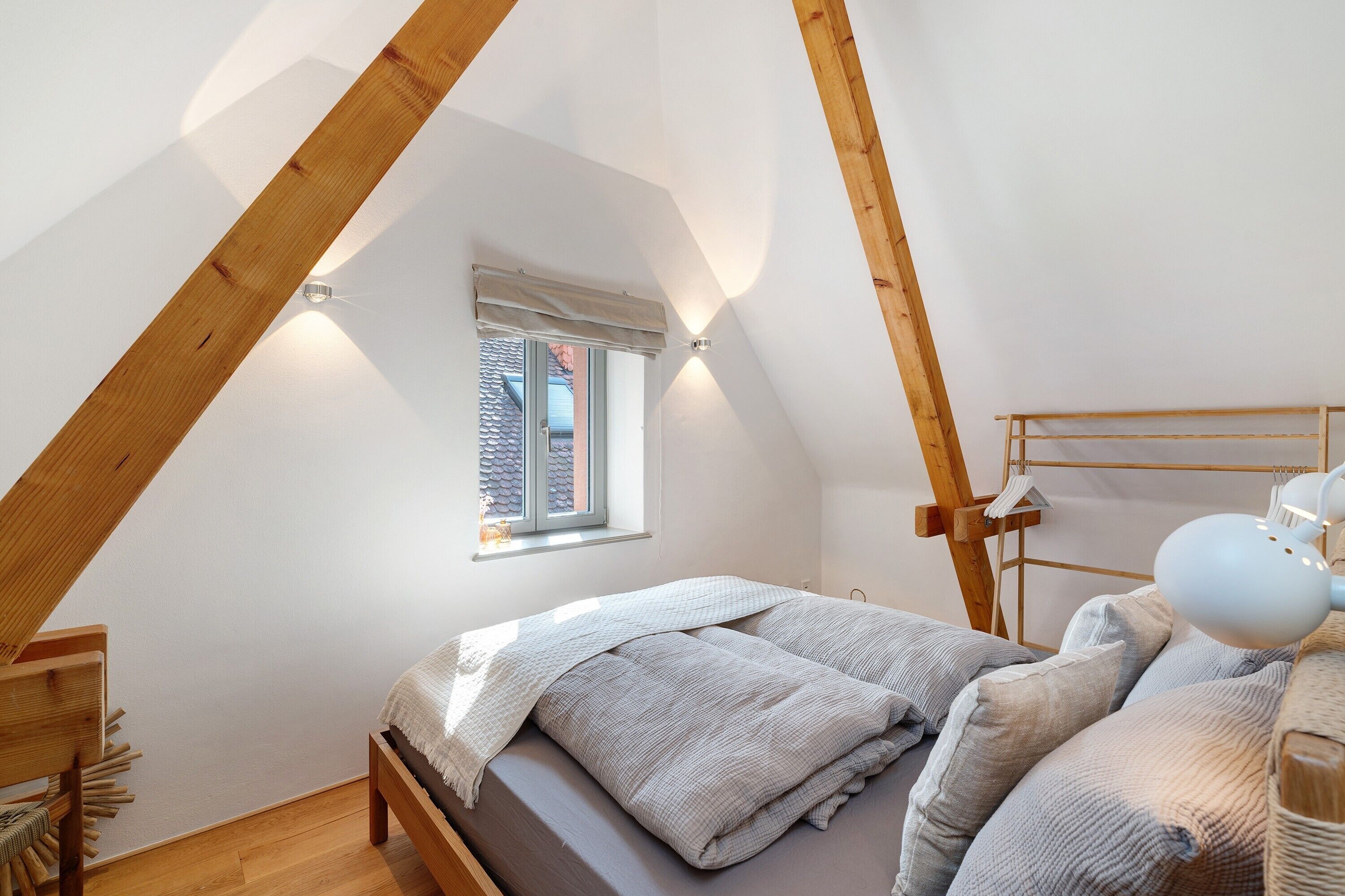 A bedroom with a sloped ceiling, a small window, a bed with pillows, a bedside lamp, and a wooden headboard.