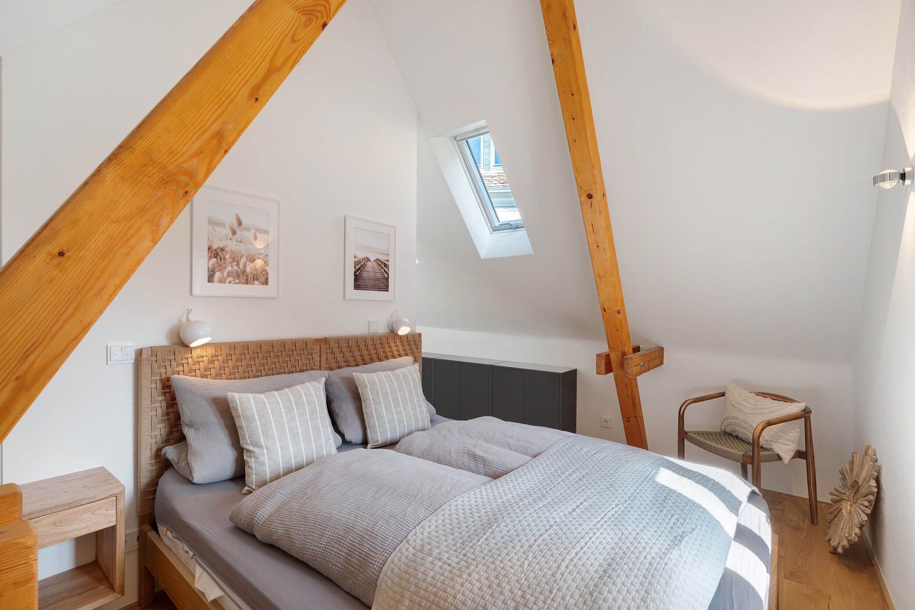 Une chambre avec un plafond en pente, un lit avec une tête de lit, une chaise et deux tableaux accrochés au mur.