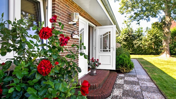 Uma casa de tijolos com porta branca, um vaso com flores vermelhas e um gramado bem cuidado.