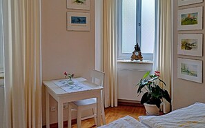 A room with a white table, a chair, a potted plant, a window with a view, and framed pictures on the wall.