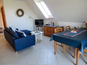 Una habitación con un sofá azul, una mesa de comedor con sillas, un televisor, un escritorio con una computadora y una ventana con vistas.