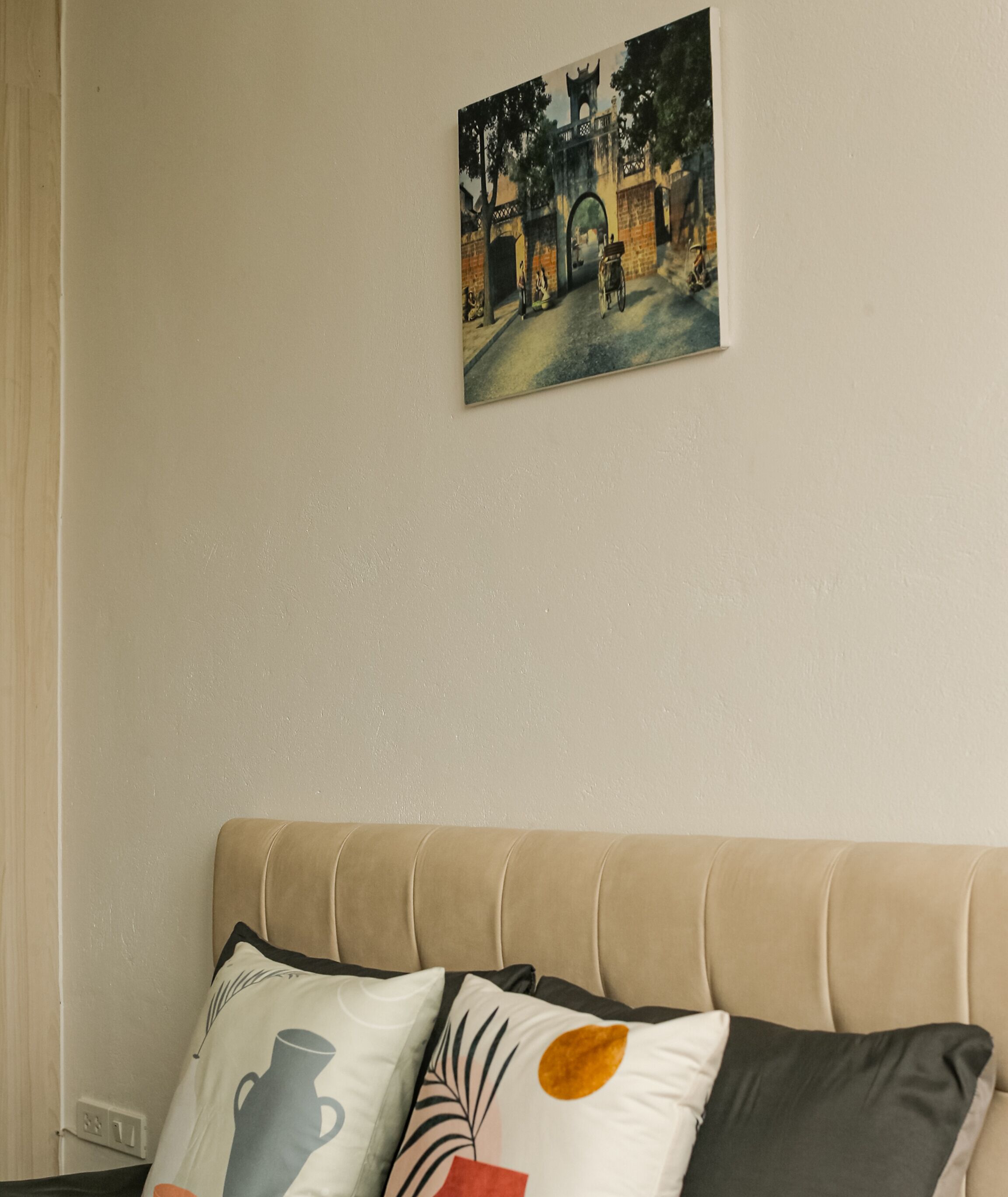 Un sofá color beige con tres cojines decorativos, un cuadro de una escena callejera y una pared lisa.