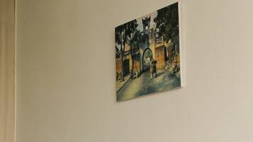 Un sofá color beige con tres cojines decorativos, un cuadro de una escena callejera y una pared lisa.