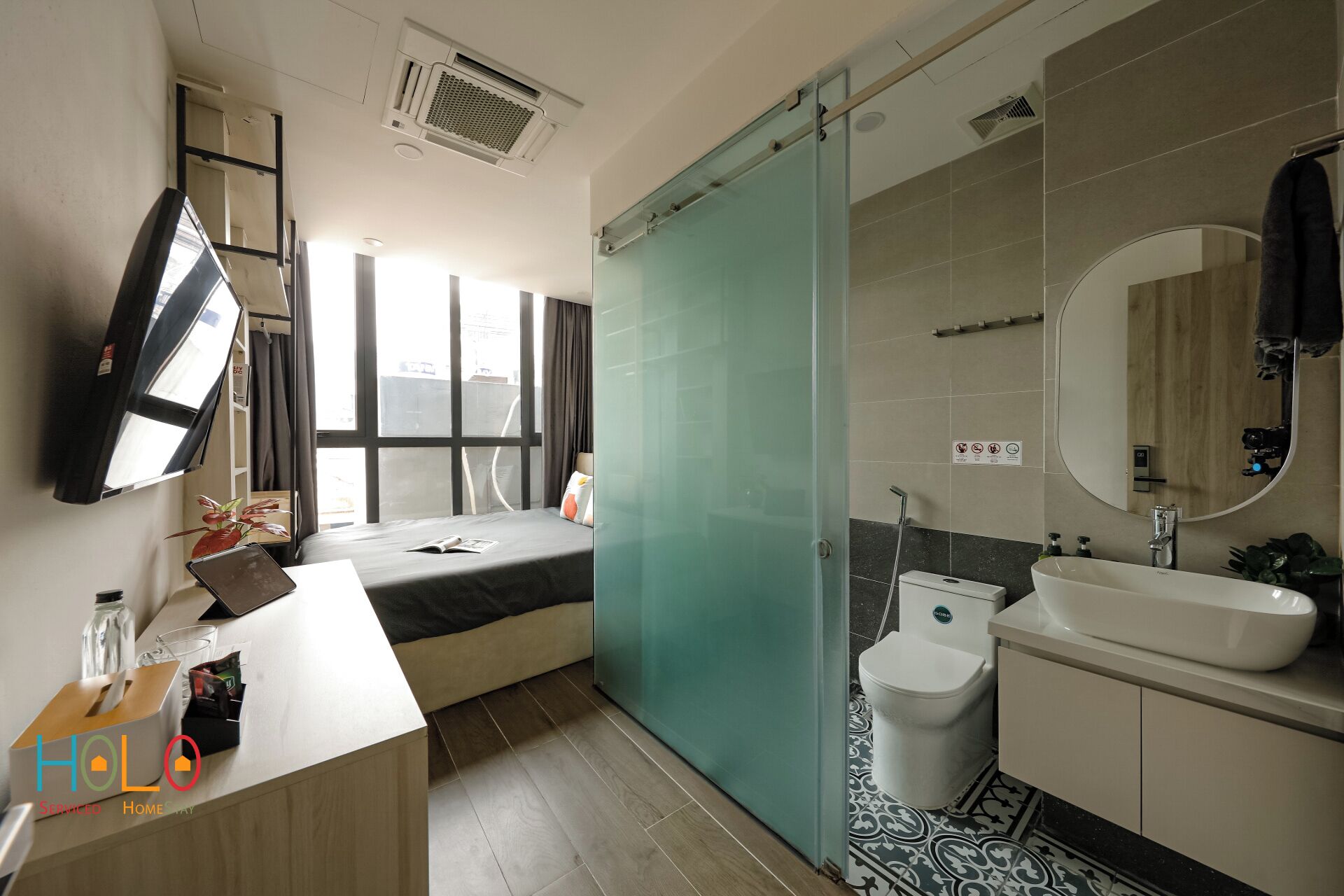A modern hotel room with a bed, a desk with a laptop, a large window, a shower area, and a bathroom with a sink and toilet.