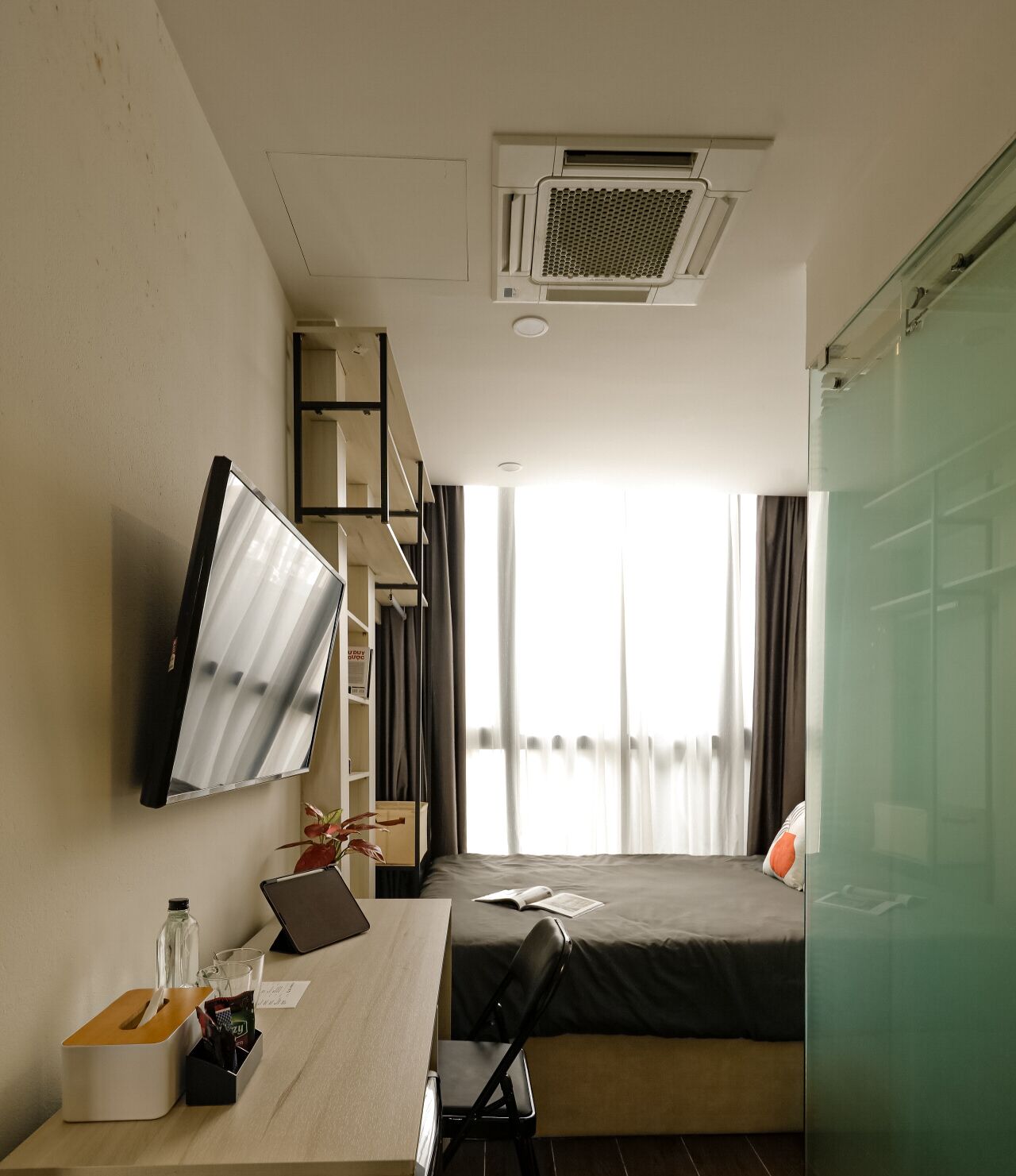 Quarto de hotel com cama, escrivaninha com computador, televisão e janela com cortinas.