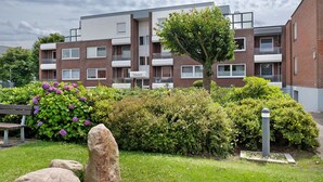 Un edificio residencial de varios pisos con balcones, un jardín con arbustos y flores, y una banca.
