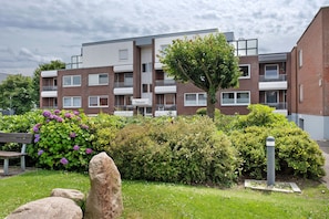 Un edificio residenziale a più piani con balconi, un giardino con arbusti e fiori, e una panchina.