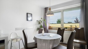 Un comedor con una mesa redonda, sillas, una lámpara colgante, una ventana con cortinas y una planta en el alféizar.