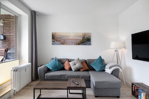 A modern living room with a grey sectional sofa, a coffee table, a mounted TV, and a painting of a sunset over water.