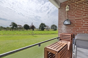 A balcony with a wooden chair, a metal railing, and a view of a green field and trees.