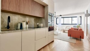 A modern kitchen with a marble countertop, wooden cabinets, and a view into a living area with a sofa and a coffee table.
