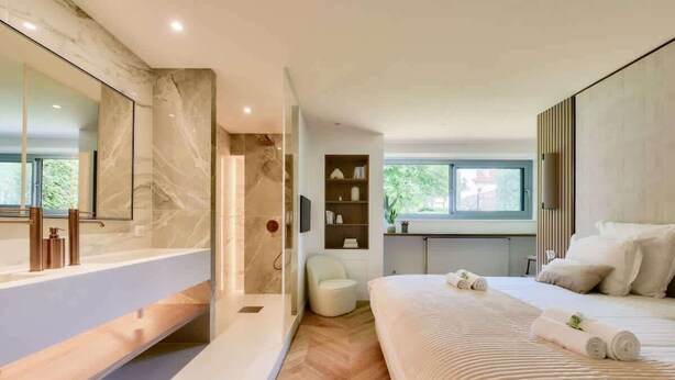 A modern bedroom with a large bed, a bathroom with a bathtub, and a view of greenery outside.