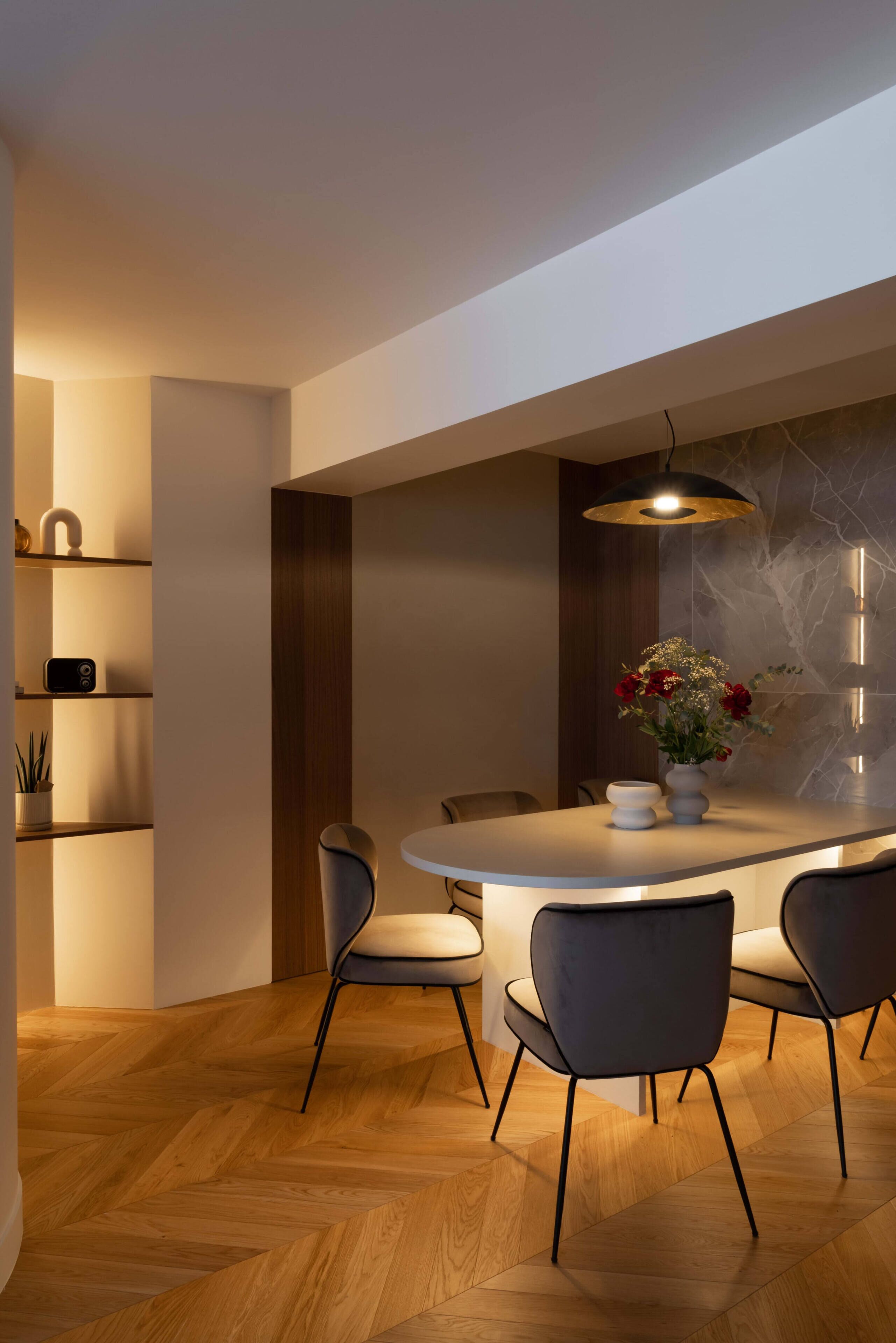 Un comedor moderno con una mesa redonda, cuatro sillas, una lámpara colgante y un jarrón con flores.