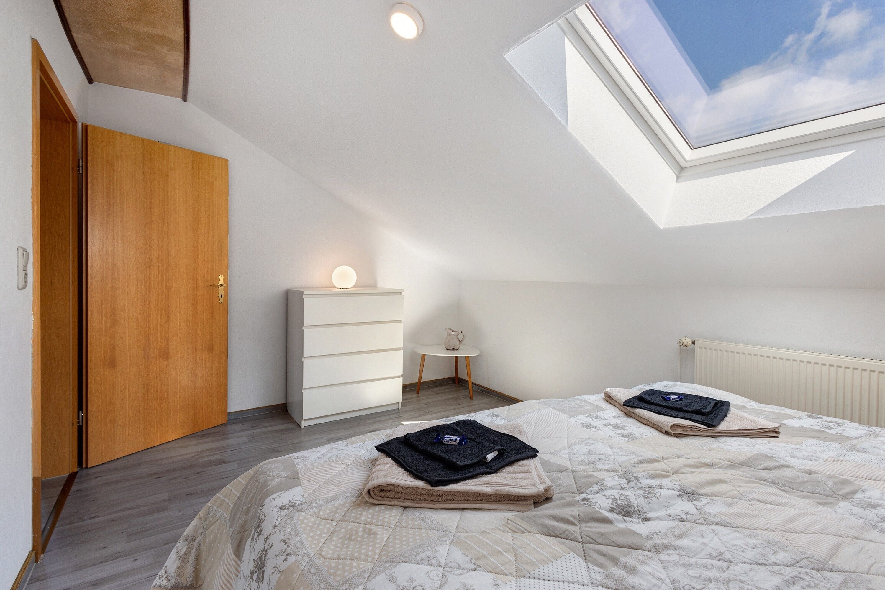 Ein Schlafzimmer mit Oberlicht, einer Holztür, einem weißen Schminktisch und einem kleinen Tisch, sowie einem Bett mit gefalteten Handtüchern.