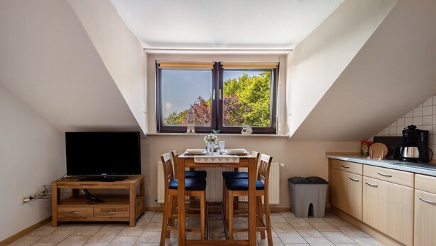 A small kitchen with a dining table, chairs, a TV on a wooden stand, a window with a view of trees, and a tiled floor. - Apartment 'Ferienwohnung Thomée' with Terrace and Wi-Fi (Looft)