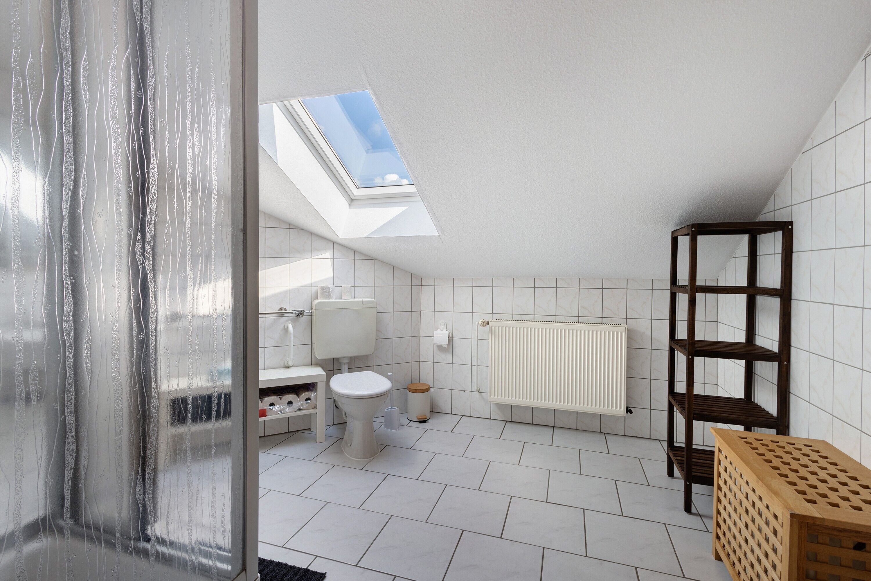 A modern bathroom with a skylight, white tiles, a toilet, a radiator, and a wooden shelf.