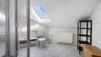 A modern bathroom with a skylight, white tiles, a toilet, a radiator, and a wooden shelf.