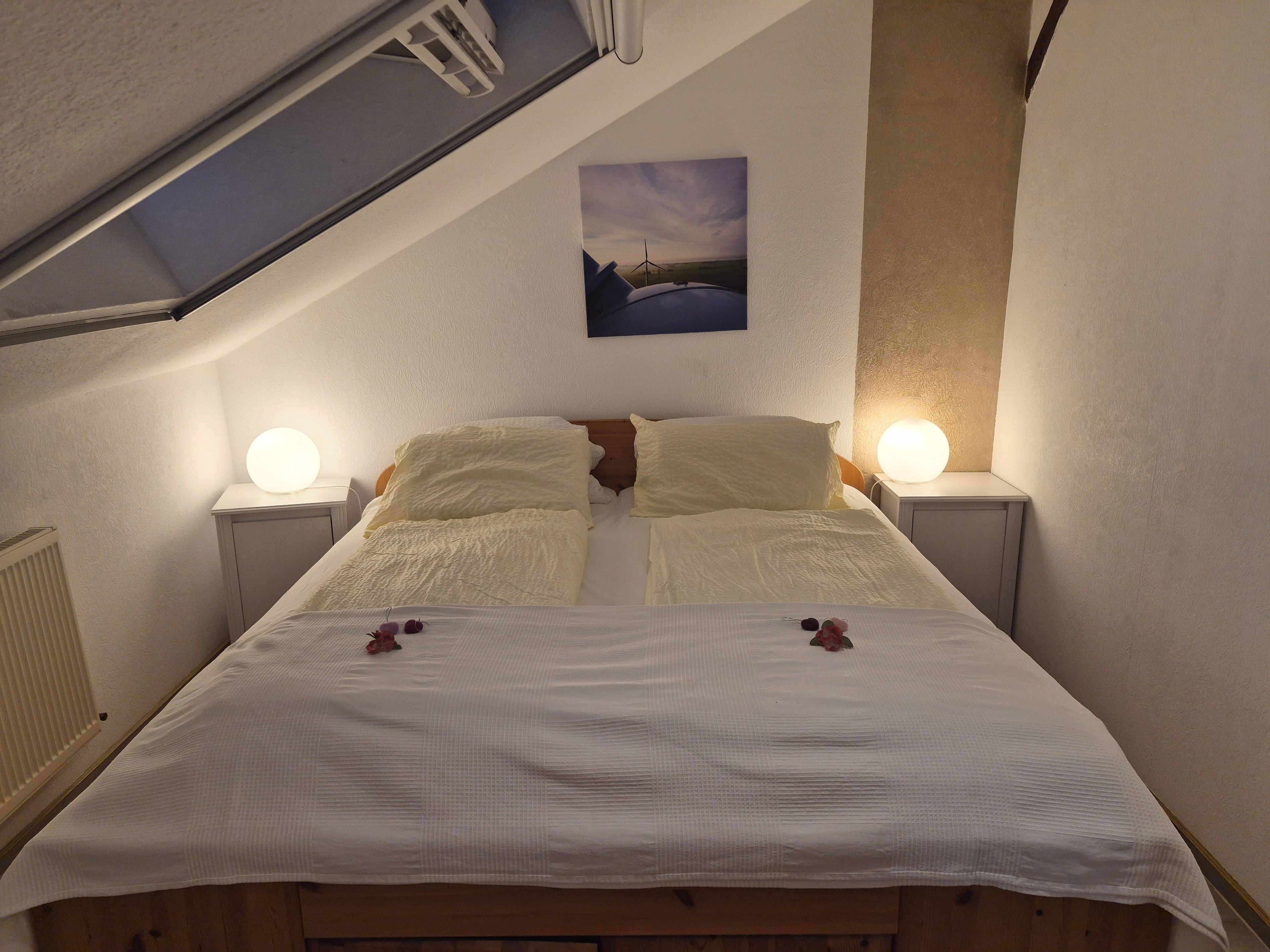 A bedroom with a sloped ceiling, two bedside lamps, a bed with a white bedspread, and a painting on the wall.