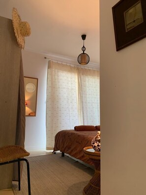 A bedroom with a bed, a chair, a hanging light fixture, a mirror, and a window with curtains.