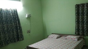 Une chambre avec un lit simple, des murs verts, des rideaux à motifs et une prise électrique visible.