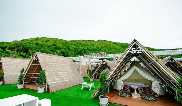 A series of thatched-roof, triangular tents with numbers indicating room assignments, set on a green lawn with a forested hill in the background. - Tawaen Caza Sky Camping (Koh Lan)