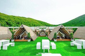 A series of triangular tents with thatched roofs, arranged in a row on a grassy field with a mountainous backdrop. - Tawaen Caza Sky Camping (Koh Lan)