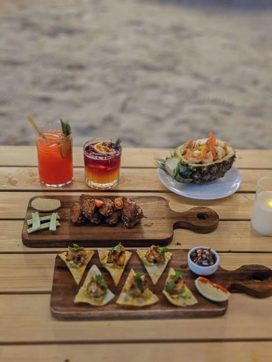 A wooden table with a variety of food and drinks, including grilled meat, appetizers, and two cocktails, set against a beach backdrop.