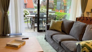 A living room with a sofa, a coffee table, and a view of a red-roofed building through a glass door. - Das Lalelou- Familienurlaub Zentral in Braunlage mit Eigenem Garten (Braunlage)