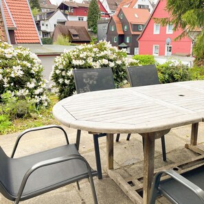 A round wooden table with metal chairs, surrounded by white flowering bushes, set on a concrete patio with houses in the background. - Das Lalelou- Familienurlaub Zentral in Braunlage mit Eigenem Garten (Braunlage)