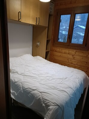 A compact room with a bed, wooden cabinets, and a window with a view of a snowy landscape. - Appartement Douillet au Calmerais un Village Typique Savoyard (Châtel)