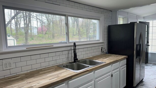 A kitchen with white cabinets, a black refrigerator, a double sink, and a window overlooking a wooded area. - Serene Country Cottage (Woodbridge)