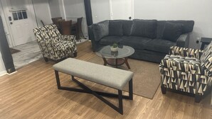 A living room with a dark grey sectional sofa, two patterned armchairs, a round coffee table with a plant, and a grey bench. - Serene Country Cottage (Woodbridge)